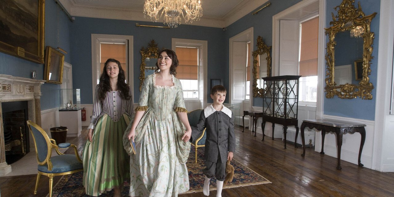 actors dressed in old garments that resemble Bridgerton. The room is decorated with b lude walls and gold accents 