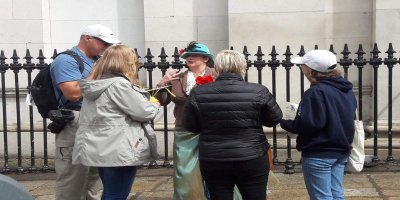 Daltons Musical Walking Tour 4 tourists standing and listening to tour guide singing. Standing infront of black gates and white building.